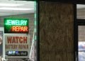 Storefront of a jewelry repair shop at night with a lit Jewelry Repair sign in the window and a boarded-up window panel beside it