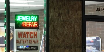 Storefront of a jewelry repair shop at night with a lit Jewelry Repair sign in the window and a boarded-up window panel beside it