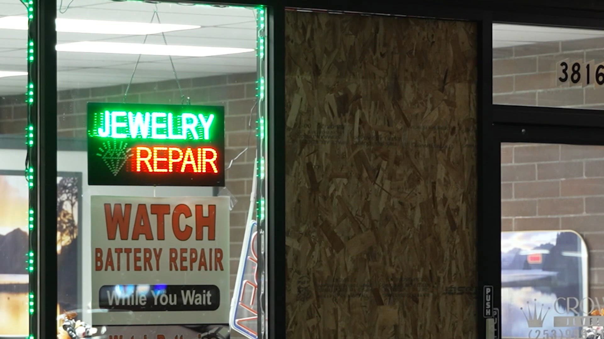 Storefront of a jewelry repair shop at night with a lit Jewelry Repair sign in the window and a boarded-up window panel beside it