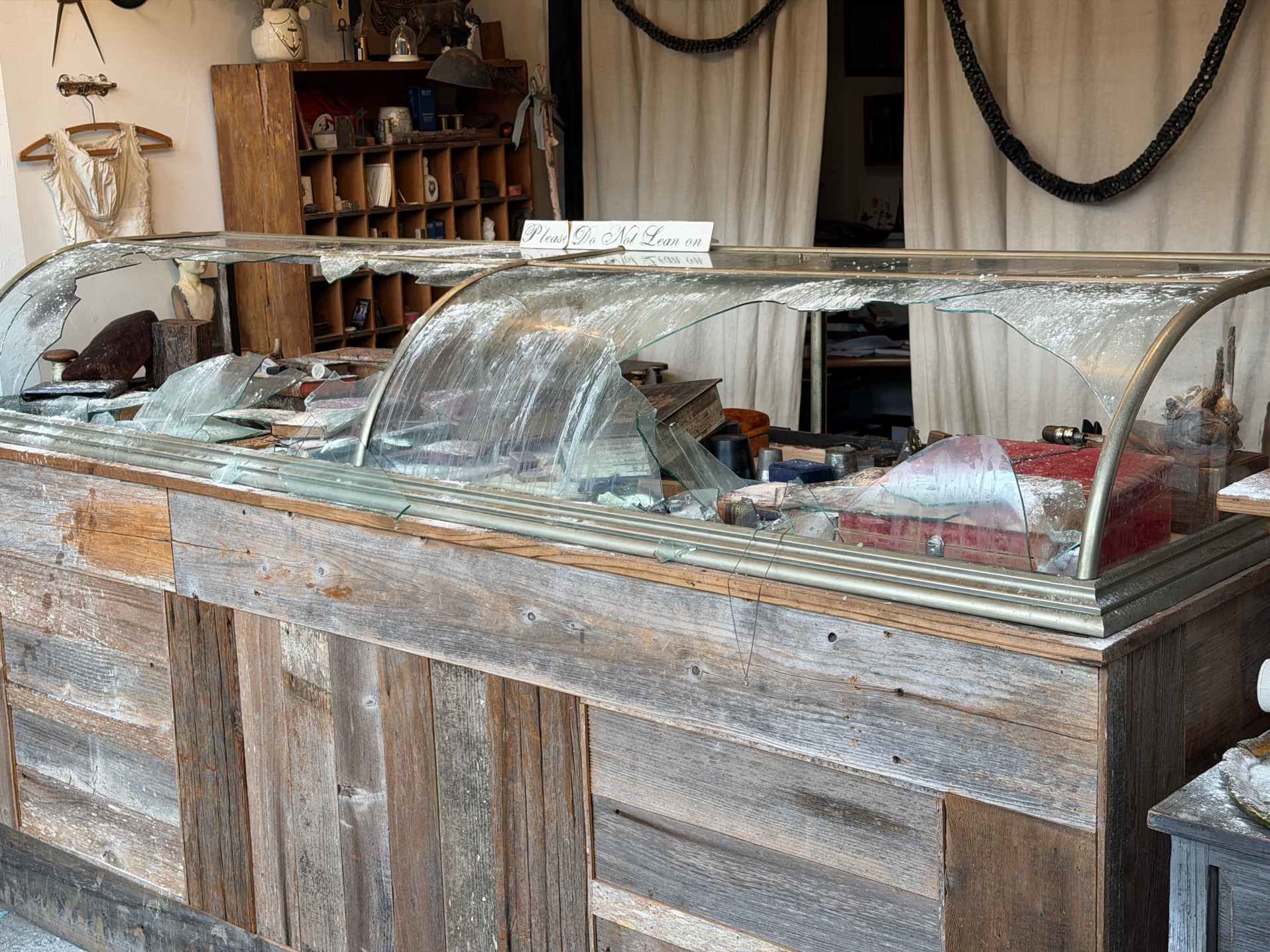 Smashed curved-glass display case on a reclaimed-wood counter inside the Modern Relics jewelry studio after the burglary